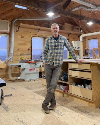 Ben Rafael, owner of Wooden Hammer, standing in his workshop. He's leaning on a table and smiling.