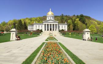 A view of the Vermont State House front with spring flowers in front and greening trees behind