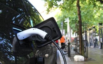 Close up of an electric vehicle charging port with charging cable attached. The car is on a city street with a sidewalk and trees in the background.